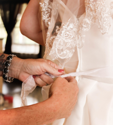 A beautiful bride smiling with anticipation as her lace dress is expertly tied in preparation for her special day.