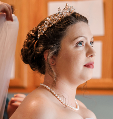 A stunning bride adorned with a golden tiara glistening in the sun, ready to have her veil placed on her head.