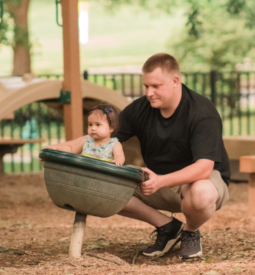 A father and daughter playing on a playground, the little girl in a colorful plastic turtle shell, and the father smiling as he watches her.