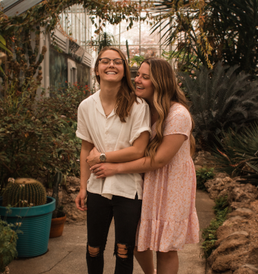Two women in love, one with a huge smile on her face as the other embraces her from behind.