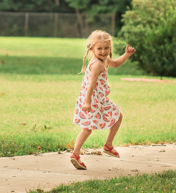 A carefree girl in a watermelon dress skipping down the sidewalk with playful pigtails bouncing.